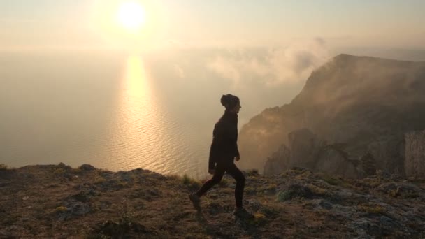 Primer plano de una joven turista en un bandana caminando a lo largo de un acantilado de un alto acantilado sobre el mar contra una hermosa puesta de sol en una cálida noche de otoño. En el fondo de la montaña hay vapor de aire . — Vídeo de stock