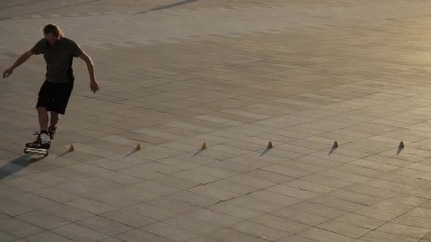 Jonge langharige man rolschaatser danst tussen kegels in een leuke avond in een stadspark. Freestyle slalom Rolschaatsen tussen kegels in slow motion. — Stockvideo
