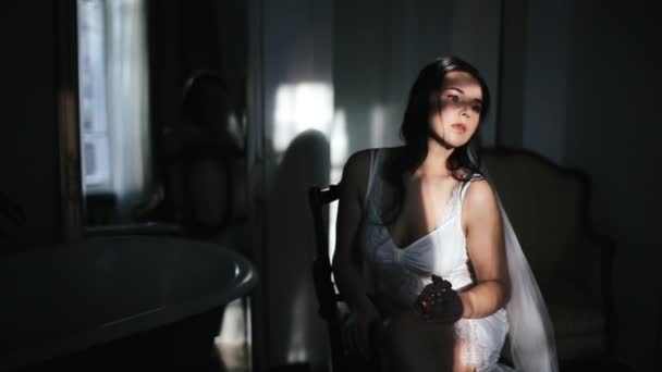 Γοητευτική νεαρή νύφη χιλιετή σε σέξι λευκό peignoir, εσώρουχα και πέπλο στο κεφάλι της κάθεται στην καρέκλα με μπουκάλι άρωμα στο σκοτεινό δωμάτιο κοντά στον καθρέφτη και το μπάνιο, ακτίνες του ήλιου πέφτουν στο σώμα από το παράθυρο — Αρχείο Βίντεο