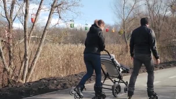 Young dad and mom are roller skating with the baby in a stroller, rear view. Sports parents roller skate on the asphalt in a warm, cozy park on a sunny day. Active hobby and good time on the street. — Stock Video