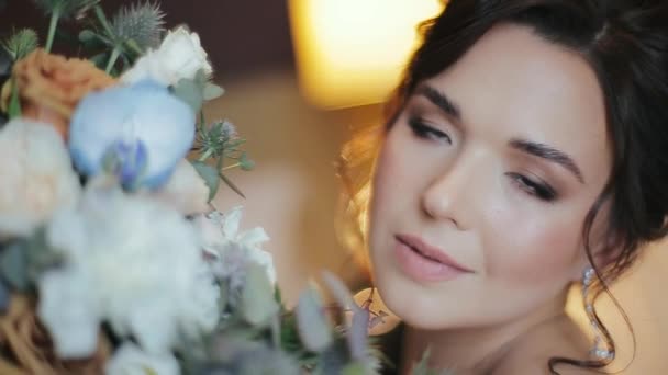 Manhã feliz de noiva millennial jovem atraente europeu com acenando olhos castanhos segurando e cheirando um luxuoso pequeno buquê de casamento de flores de rosas multi-coloridas contra a luz da lâmpada brilhante . — Vídeo de Stock