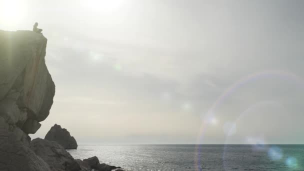Silhueta de turista sentado em cima de um penhasco contra um pano de fundo de brilho do sol e do mar. Vida ativa de uma pessoa que adora caminhadas e camping. O homem desfruta da paisagem do mar e do céu no verão . — Vídeo de Stock