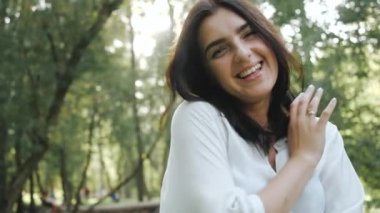 Güzel bir fotoğraf için oldukça olumlu esmer şirin gülümsemeler. Mutlu genç kadın, öğleden sonra güzel bir fotoğraf için poz veriyor. Arkalarında bahar ağaçları ve parlak güneş var..