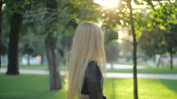 Mooi Kaukasisch meisje mode model alleen in het park, draait en glimlacht, kijkt naar de camera, loopt en straalt haar lange blonde haar. Jonge vrouw die buiten poseert. Mensen en stijl concept. — Stockvideo