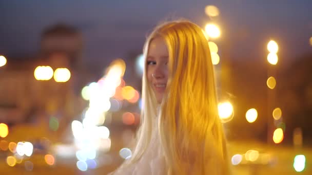 Hermosa modelo de moda chica caucásica caminando en la ciudad nocturna, feliz y sonriente. Una joven maquillada con un vestido elegante endereza su largo cabello rubio y posa sobre un fondo de luces . — Vídeos de Stock