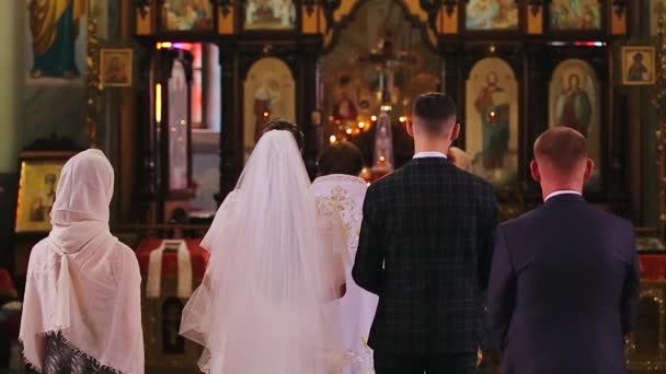 Vue arrière de la cérémonie chrétienne de mariage de la mariée dans une belle robe blanche et le marié dans un costume et deux témoins debout à proximité. Le prêtre couronne jeune couple de mariage à l'autel . — Video