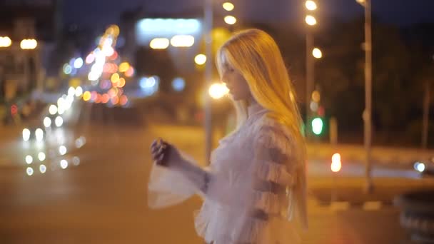 Hermosa modelo de moda chica caucásica caminando por la ciudad nocturna, feliz y sonriente. Mujer joven con maquillaje en un vestido elegante blanco endereza su cabello largo y posa contra el telón de fondo de las luces . — Vídeos de Stock