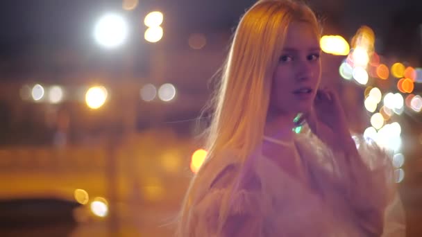 Девушка-модель прогуливается по ночному городу, счастливая и улыбающаяся. Молодая женщина с макияжем в стильном платье выпрямляет длинные светлые волосы и позирует на фоне огней — стоковое видео