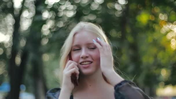 Beautiful caucasian girl fashion model walks in nature and speaks on the phone, smiles and straightens her long blonde hair. Young stylish quarantined woman outdoors in sun. Aesthetic people concept — Stock Video
