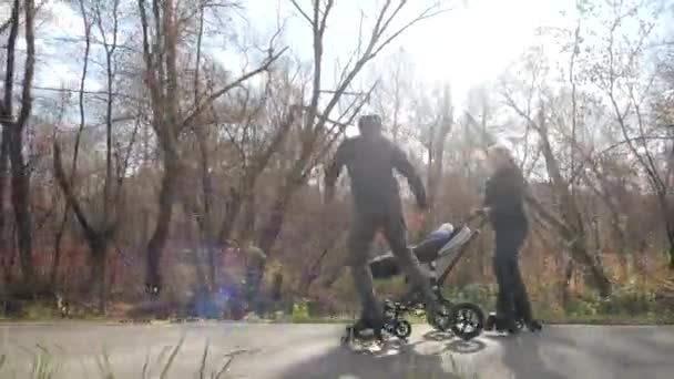 Jóvenes padres alegres montados en patines con un bebé en un cochecito a lo largo del callejón de la ciudad bajo el sol. Papá da un salto y entretiene al niño. Movimiento lento . — Vídeos de Stock