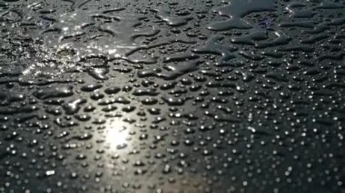 Parlak, pürüzsüz zemine damlayan hafif yağmur birikintileri, yakın görüş. Güneşin su damlalarındaki yansıması. Güneşli ve yağmurlu hava. Su grafit renginin yüzeyine dökülür..