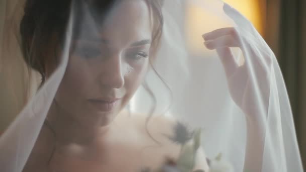 Close-up uitzicht op een charmante mooie blanke bruid in een luxe witte trouwjurk en sluier met een bruidsboeket bloemen achter een sluier in de gezellige slaapkamer van een hotelkamer in de ochtend. — Stockvideo