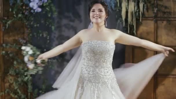 Feliz encantadora novia milenaria morena en velo y vestido de novia decorado con perlas y diamantes de imitación sostiene un ramo de flores en sus manos y remolinos contra el fondo de la puerta y la planta — Vídeos de Stock