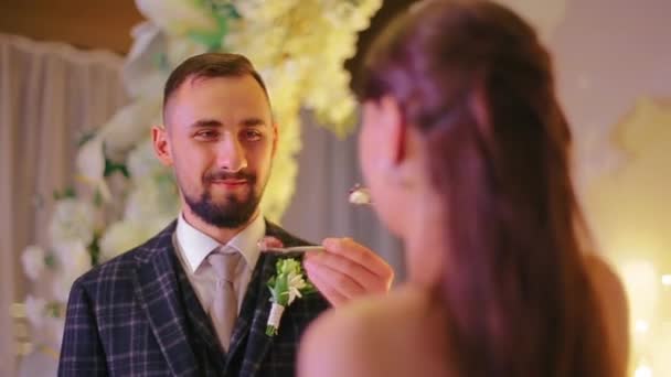 Close-up view of young wedding couple eating delicious and mouth-watering cake on brudershaft. The bride and groom treat each other with cake from spoon on the background of altar in the banquet hall. — Stock Video