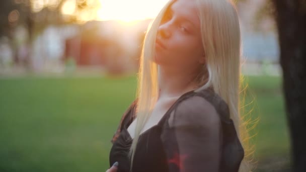 Joven chica caucásica, una hermosa modelo de moda en la naturaleza, inclina la cabeza y endereza el cabello largo y rubio. Adolescente estética en un vestido negro con manicura en el parque en los rayos del sol poniente — Vídeo de stock