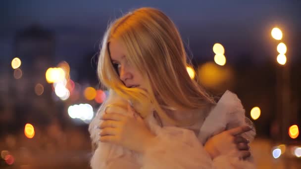 Aantrekkelijk en teder Kaukasisch tienermeisje knuffelt zichzelf 's nachts in de stad op een achtergrond van licht. Jonge mode model met lang blond haar in een jurk poseren. Esthetiek en trends. — Stockvideo