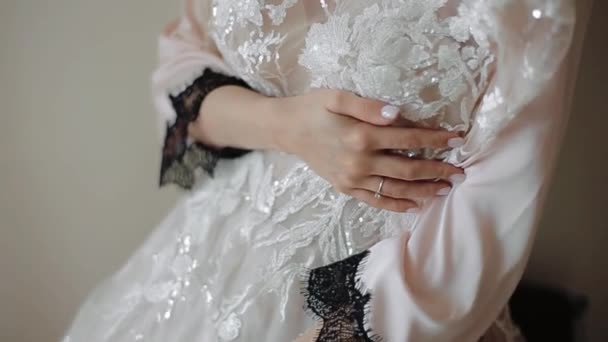 Una giovane bella sposa in una stanza luminosa in un negligee stringe un abito da sposa bianco con ricami in pizzo lucido con le mani con una manicure e un anello di fidanzamento — Video Stock