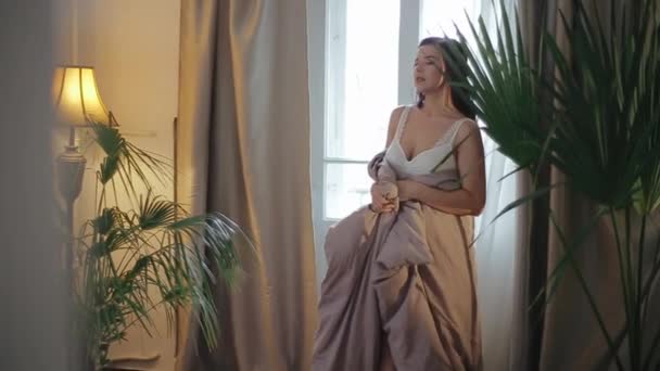 Mooie jonge Kaukasische brunette bruid in luxe witte lingerie bedekt met deken wandelingen rond hotelkamer en geniet 's morgens op de achtergrond van indoor palm bloemen, lichtgevende vloerlamp. — Stockvideo