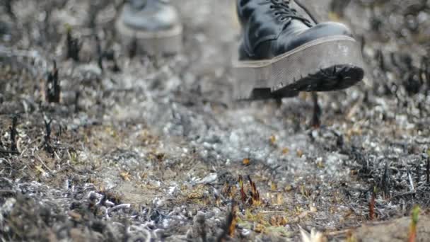 Fille moderne en cuir à la mode bottes noires avec semelles hautes marche le long de la cendre brûlée de l'herbe dans un champ inhabité et s'arrête, se concentrant sur ses pieds avec des chaussures, vue du bas au ralenti . — Video