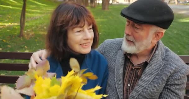 Весела пара людей похилого віку чоловік і жінка сидять на лавці, відпочиваючи в затишному парку восени. Бородатий красивий літній чоловік жартує, обіймає і цілує свою дружину букетом листя . — стокове відео