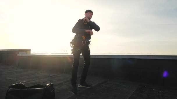 Professional sports industrial climber stands on the roof and fixes various cable trailers on his body to go downstairs outside the house, against background of the sun, close-up view. Dangerous job. — Stock Video