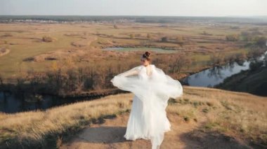 Güzel milenyum gelini çimenlerin arasında bir tepede duruyor ve muhteşem duvak gelinliğini dışarıda güneşli bir günde, sonbahar manzarasının, tarlaların ve nehirlerin arka planına sallıyor..