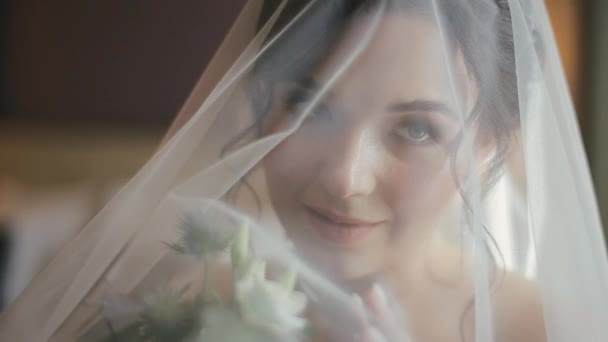 Close-up shot van aantrekkelijk gezicht van aantrekkelijke jonge bruid in een sluier en witte trouwjurk staande in hotelkamer met boeket van verse mooie bloemen in de ochtend. Gelukkige vrouw voor de bruiloft. — Stockvideo