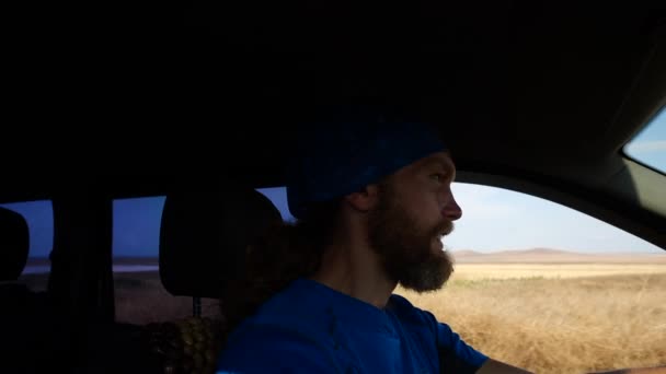 Primer plano de turista barbudo en bandana azul y camiseta sentada al volante de un coche y conduciendo en la carretera en medio del campo en un día soleado y caluroso, luego saludando a alguien y riendo . — Vídeos de Stock