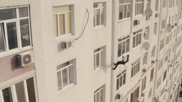 Plano superior de escalador industrial masculino en cable con ramo de rosas colgando en la pared del edificio de apartamentos en el exterior, trabajando como mensajero para entregar flores a la altura que desciende cuidadosamente por la cuerda — Vídeos de Stock