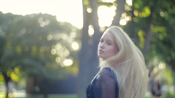 Ein junges attraktives kaukasisches Mädchen dreht sich um, blickt in die Kamera und richtet ihre langen blonden, gesunden Haare. Schickes Teenie-Model lächelt selbstbewusst und posiert im Park. — Stockvideo