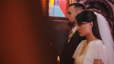 Kilisede birbirine aşık iki kişinin Hıristiyan düğün töreninin yan görüntüsü. Bulanık duvar lensin zeminini kaplıyor. Genç inançlı gelin ve damadın ellerinde mumlarla yan görünümü.
