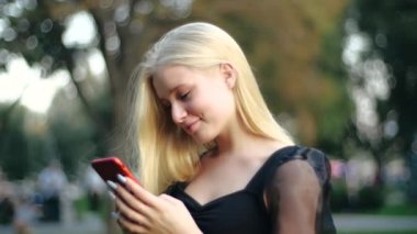 Siyah bahar elbisesi içinde güzel, gülümseyen genç bir sarışın elinde kırmızı bir akıllı telefon tutuyor ve dışarıda ağaçların arasında sıcak, rahat şehir parkında arkadaşlarıyla sohbet ediyor. Telefonu olan kız..