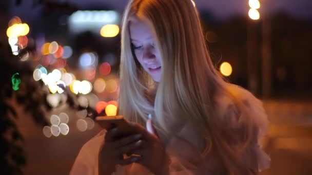 Niedlich lächelnde und glückliche Blondine mit langer Maniküre und im weißen Kleid, die mit Freunden auf dem Smartphone vor dem Hintergrund verschwommener Lichter von Autos und Bäumen im Zentrum der Abendstadt plaudert, Nahaufnahme. — Stockvideo