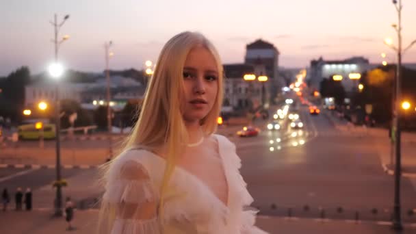 Menina loira maravilhosa bonita com cabelos longos e vestido de luz branca posa na câmera no centro da cidade contra fundo da estrada, carros itinerantes e luzes de rua da cidade noturna de Kharkov, Ucrânia. — Vídeo de Stock