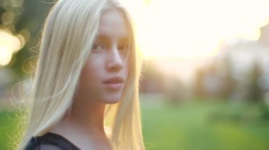 Beyaz, uzun saçlı, baharda çimlerin ve ağaçların arka planında neşeli bakışlı güzel bir genç kızın parkında yürümek, yavaş çekimde yakın çekim. Çekici, güzel sarışın dışarıda dinlenmeyi seviyor.
