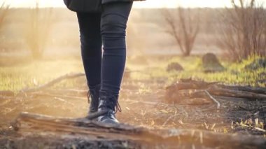 Genç bir kızın önünde sinematik çekimler moda taytlar ve şık siyah yüksek tabanlı çizmelerle gün batımının altındaki bir tarlaya ateş üstüne ateş. Baharda yanan çimlere basan kişi.
