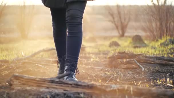 Cinematic shot voor jong meisje wandelen in trendy leggings en stijlvolle zwarte hoge zool laarzen op as grond na vuur tegen een veld onder zonsondergang. Persoon stapt in het voorjaar op verbrand gras. — Stockvideo