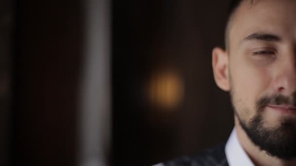 Close-up view of half face of millennial bearded European man in white shirt with a plaid vest. Stylish bearded caucasian groom smiles and looks at camera against the background of a dark hotel room — Stock Video