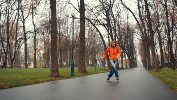 Turuncu ceketli ve kot pantolonlu aktif sakallı profesyonel patenci Gorky Park 'ın asfalt yolu boyunca ağaçlara ve yürüyen insanlara karşı bahar yağmurundan sonra ustaca kayıyor.. — Stok video