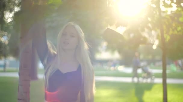 Jovem caucasiana com longos cabelos loiros ao sol no parque da cidade toca folhas verdes com a mão. Modelo adolescente na blusa preta casual elegante vira-se, sorri docemente, posa. Moda e ecologia . — Vídeo de Stock