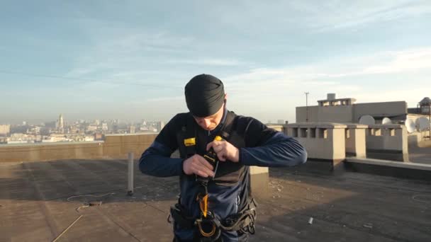 Erkek bir endüstriyel tırmanıcının çatıda dikilip sıcak bahar havasında vücuduna kanca taktığı ve kablo bağladığı yakın çekim. Tehlikeli ve aşırı riskli bir iş.. — Stok video
