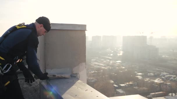 Fechar tiro de alpinista profissional do sexo masculino em uniforme, de pé no telhado contra o pano de fundo do sol brilhante, fixação e fixação de cabo de segurança para descer ao longo da parede da casa. Desporto extremo . — Vídeo de Stock