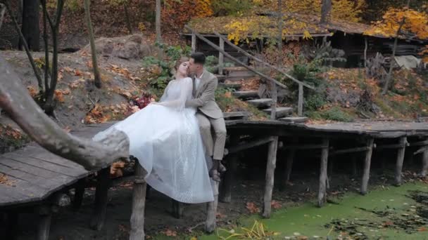 Casal jovem emocional sensível sentado em pequena ponte de madeira por lago pantanoso com olhos fechados, desfrutando de solidão na bela natureza pela floresta de outono. Noiva apoiada no noivo na ponte. — Vídeo de Stock