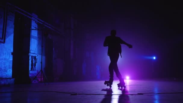 Prise de vue arrière d'un patineur professionnel portant un bonnet et une chemise autour de la scène sous un éclairage bleu et violet et effectuant divers tours et tours complexes. Performance expérimentée du patineur à roulettes — Video