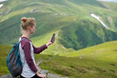 Onu telefona arıyorsunuz bir kayaya oturan genç bir kadın. Arka planda dağların muhteşem görünümü.