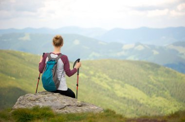Kısa mola bir yürüyüş gezisi Dağları nın nefes kesen manzarasının tadını çıkarın. Uzak kameradan bakan genç kadın bir kayaya oturan; Onu geri ve tutan trekking sopa üzerinde küçük sırt çantası.