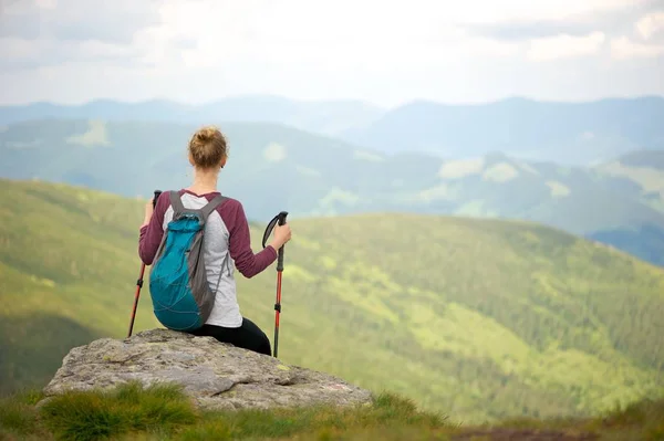 Kısa mola bir yürüyüş gezisi Dağları nın nefes kesen manzarasının tadını çıkarın. Uzak kameradan bakan genç kadın bir kayaya oturan; Onu geri ve tutan trekking sopa üzerinde küçük sırt çantası.