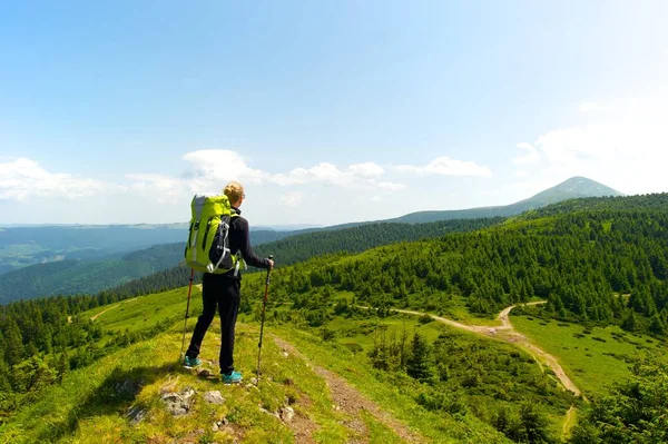 Active lifestyle concept. A young woman in black with bright green backpack and trekking sticks standing on top of a hill, taking in the amazing view of the mountains, forest and sky.