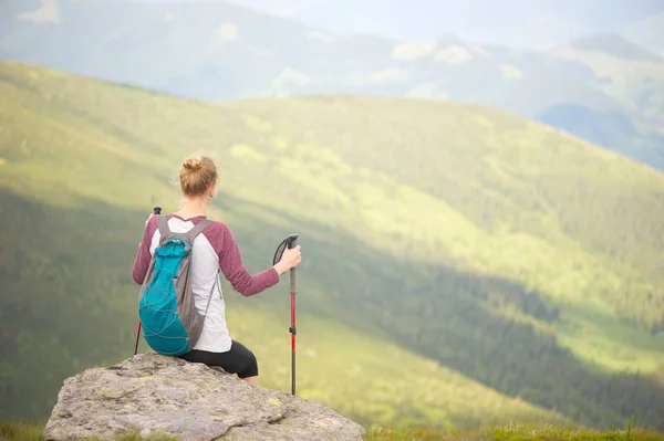 A young woman sitting on a rock taking in the amazing view of the mountains. Holding trekking sticks; backpack on her back.