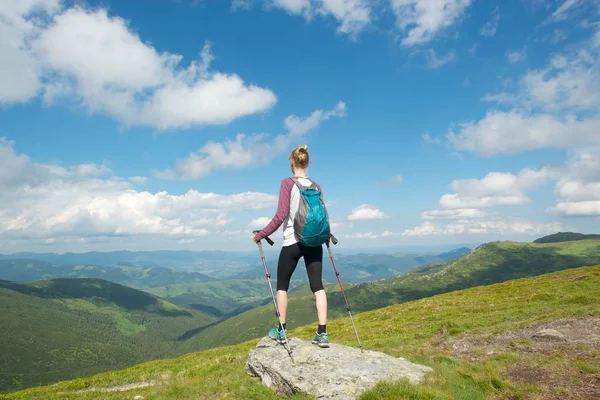 Active lifestyle concept. A young woman with a small backpack, standing on top of a hill, holding trekking sticks and enjoying an amazing view of the mountains below.